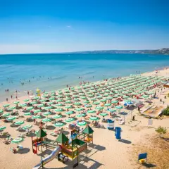 Słoneczna plaża w Złote Piaski pełna leżaków i parasoli, z ludźmi kąpiącymi się w morzu i bawiącymi na placu zabaw.