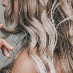 Dłoń dotyka falujących włosów w odcieniach ombre blond, tworząc subtelny efekt.