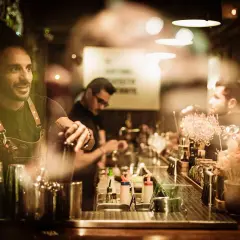 Barman sorridente prepara cocktail dietro un bancone illuminato, perfetto per sapere dove bere a Barcellona.