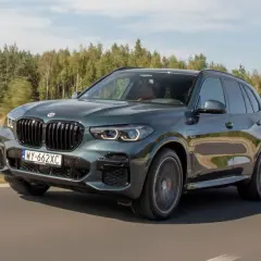 Ciemnozielone BMW X5 45e pędzi po drodze, otoczone zielenią lasów.