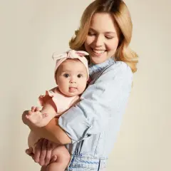 Uśmiechnięta mama tuli niemowlę w różowym body i opasce. Wskazówka, jak ubierać noworodka jesienią, by było mu ciepło i wygodnie.