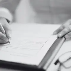 Dłoń pisze na dokumencie, obok okulary. Dowiedz się, jak sprawdzić pozwolenie na budowę sąsiada, analizując formalności.