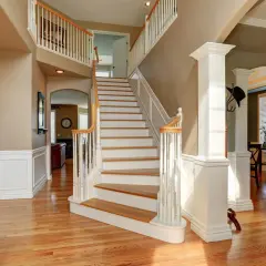 A grand foyer with a sweeping staircase featuring white painted risers and wooden treads. The staircase leads to an upper landing, with a view into a living area.