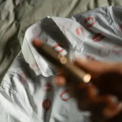 Ein weißes Hemd mit vielen roten Lippenstiftabdrücken. Eine Hand hält einen Lippenstift, um die Flecken zu entfernen.