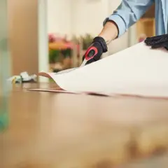 Osoba w rękawiczkach tnie papier, przygotowując go do przyklejenia. Zastanawiasz się, czym podkleić tapetę?