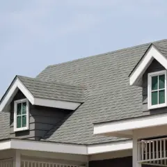 A close-up of a house roof showcasing different types of shingles in a grey hue, with dormer windows peeking out.