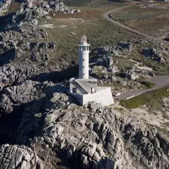 Faro blanco en una punta rocosa, con una carretera serpenteante que sube hacia él.