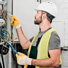 Elektryk w kasku i kamizelce odblaskowej pracuje przy skrzynce z przewodami. Zastanawiasz się, ile kosztuje elektryk?