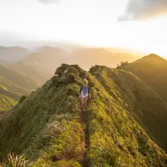 Une femme court sur un sentier de montagne verdoyant au coucher du soleil, profitant d'un parcours spectaculaire.