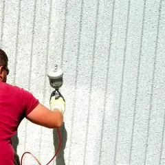Uomo con maglietta rossa usa uno spruzzatore per verniciare una parete esterna. Ottimi trucchi per verniciare a spruzzo per un lavoro uniforme.