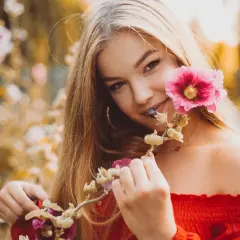 Uśmiechnięta blondynka w czerwonej bluzce podczas sesji kobiecej w plenerze, trzymająca gałązkę z różowymi kwiatami malwy.