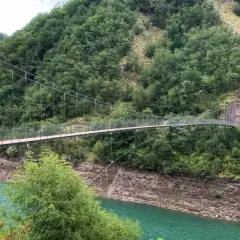 Un ponte sospeso attraversa un lago turchese, circondato da montagne boscose. Due persone passeggiano sul ponte, godendosi il panorama dei vagli di sotto.