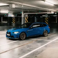 Niebieskie BMW M3 Touring z czarnymi pasami na masce stoi na pustym parkingu podziemnym.