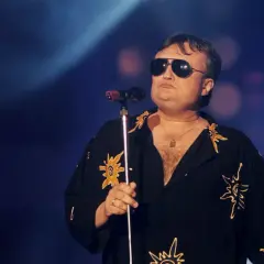 Wokalista Budki Suflera w okularach przeciwsłonecznych śpiewa do mikrofonu. Jego koszula ozdobiona jest słońcami.