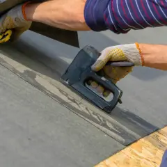 Ręce w rękawiczkach mocują rolkę papy za pomocą zszywacza.