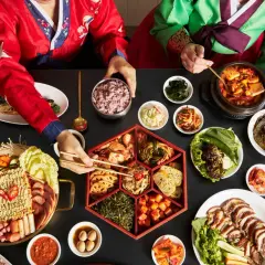 Dwie osoby w tradycyjnych koreańskich strojach jedzą obfity posiłek z wieloma daniami, w tym kimchi.