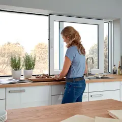 Kobieta przygotowuje jedzenie w jasnej kuchni z dużymi oknami. W tle widać drzewa, a na blacie stoją zioła i akcesoria kuchenne.
