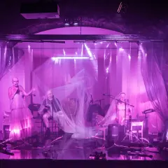 Na scenie teatru ludowego, w fioletowym świetle, artyści prezentują swój repertuar. Wokalistka w zwiewnej sukni i muzycy tworzą magiczną atmosferę.