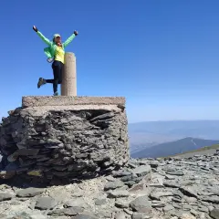 Mujer con el chullo puesto, brazos en alto, celebra en la cima de una montaña rocosa bajo un cielo azul despejado.