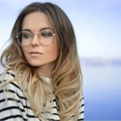 Blondynka w okularach o okrągłych oprawkach, w bluzie w paski, na tle błękitnego nieba i wody.