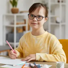 Dziewczynka z trisomią 21, w okularach, z uśmiechem rysuje kredką.