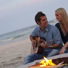 Coppia sulla spiaggia al tramonto, lui suona la chitarra, lei lo guarda sorridendo. Un falò illumina la scena, ma la noia nella coppia è palpabile.