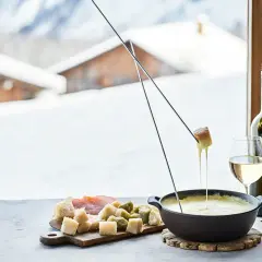 Une délicieuse recette fondue au fromage, avec du pain et des cornichons, servie avec du vin blanc devant un paysage enneigé.