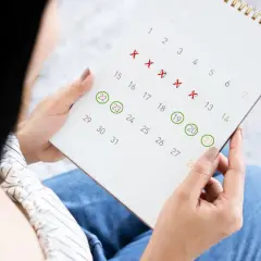 Kobieta analizuje kalendarz, zaznaczając dni. Czerwone krzyżyki sugerują pytania o to, jak nie mieć okresu, a zielone kółka oznaczają inne ważne daty.