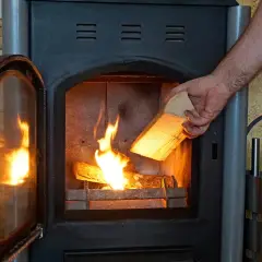 Hand legt Holzscheit in brennenden Kaminofen. Beachten Sie die Kaminofen Verordnung für sicheres Heizen.