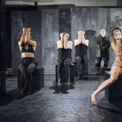 Scena z "Teatru Pieśni Kozła". Aktorzy w mrocznej scenerii, niektórzy z rękami przy twarzy, inni w krwawych strojach.
