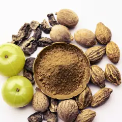 Triphala Pulver in einer Schale, umgeben von Amla-Früchten und getrockneten Haritaki- und Bibhitaki-Samen. Die Triphala Wirkung wird durch diese Zutaten erzielt.