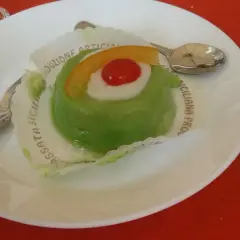 Zielone lody cassate, ozdobione wiśnią i skórką pomarańczy, na białym talerzyku z łyżeczką.