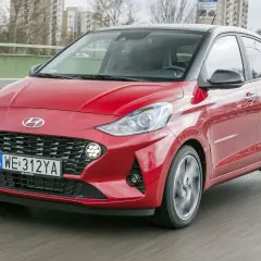Czerwony Hyundai i10 z czarnym dachem pędzi po drodze. W tle widać budynki i drzewa.
