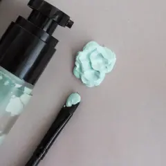 Butelka z zielonym korektorem, pędzel z produktem i plama korektora na jasnym tle.