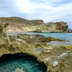 Agua turquesa en una cueva natural en la playa del carbón, con rocas volcánicas y montañas áridas al fondo.