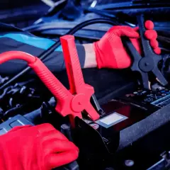 Ręce w czerwonych rękawiczkach podłączają kable rozruchowe do akumulatora. Czy nowy akumulator trzeba ładować? To pytanie często zadawane przez kierowców.