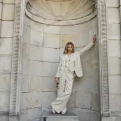 Elegancka kobieta w beżowym garniturze z żakardowym wzorem, oparta o kamienną ścianę.