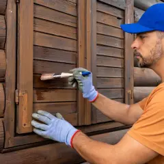 Uomo con cappellino blu e guanti blu effettua la verniciatura persiane in legno scuro, rinnovando la facciata di una casa in tronchi.