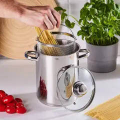 Ręka wkłada spaghetti do garnka, by ugotować je idealnie al dente. Obok pomidorki i świeża bazylia.