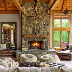 A cozy living room with a stone fireplace, antler chandelier, and leather armchairs embodies traditional rustic interior design.