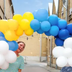 Två glada kvinnor firar en studentfest med en båge av blå, gula och vita ballonger.