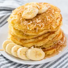 Stos puszystych naleśników z mąki kokosowej, udekorowany plasterkami banana i wiórkami kokosowymi, polany syropem.