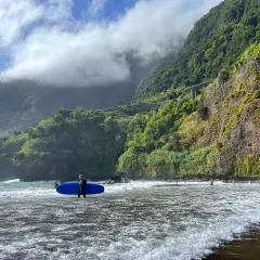 Surfer z niebieską deską na czarnej plaży Madera, w tle zielone klify i chmury.