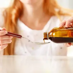 Kobieta nalewa łyżeczkę syropu z brzozy na kaszel z brązowej buteleczki.