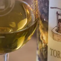 Calice di vino bianco dorato accanto a una bottiglia con etichetta che raffigura un torchio e grappoli d'uva, evocando il sapore del torchiato di fregona.