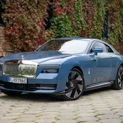 Niebieski Rolls-Royce Spectre, luksusowy samochód elektryczny, zaparkowany przed ceglanym budynkiem porośniętym jesiennymi liśćmi.