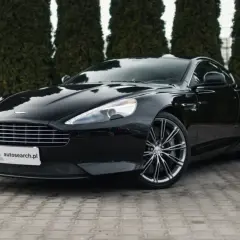 Czarny Aston Martin DB9 lśni na tle zielonych żywotników. Elegancki, sportowy samochód z charakterystycznym grillem i felgami.