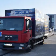 Czerwona ciężarówka MAN z naczepą WORD i literą L na masce, na placu manewrowym.