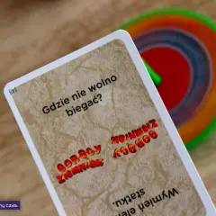 Karta z gry "Gorący ziemniak" pyta "Gdzie nie wolno biegać?". W tle kolorowy bączek.