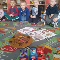 Dzieci w przedszkolu siedzą na dywanie z zabawkami, przed nimi talerz z kawałkami czekolady i opakowania czekolad.